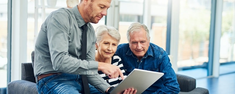 Plan d'un couple de personnes âgées utilisant une tablette numérique lors d'une consultation avec un conseiller financier à domicile.