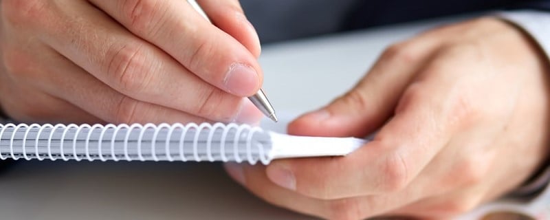 Homme d'affaires tenant un stylo et écrivant sur un bloc-notes avec des pièces de monnaie et une calculatrice à côté.