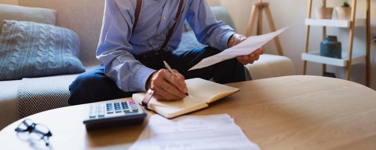 Un homme âgé de race blanche qui examine ses finances à la maison et analyse les factures tout en écrivant dans un agenda.