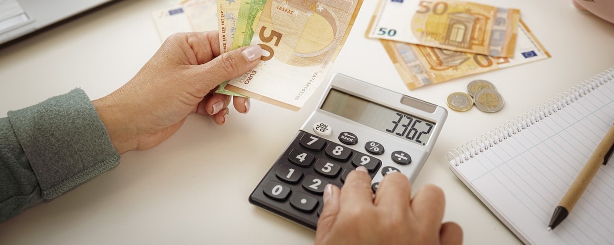 Gros plan des mains d'une femme utilisant une calculatrice pour planifier les finances de son foyer. Elle tient des billets de banque en euros.