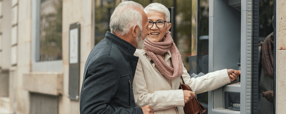 De joyeuses personnes âgées aux cheveux gris avec des sacs en papier retirant de l'argent à un distributeur automatique.