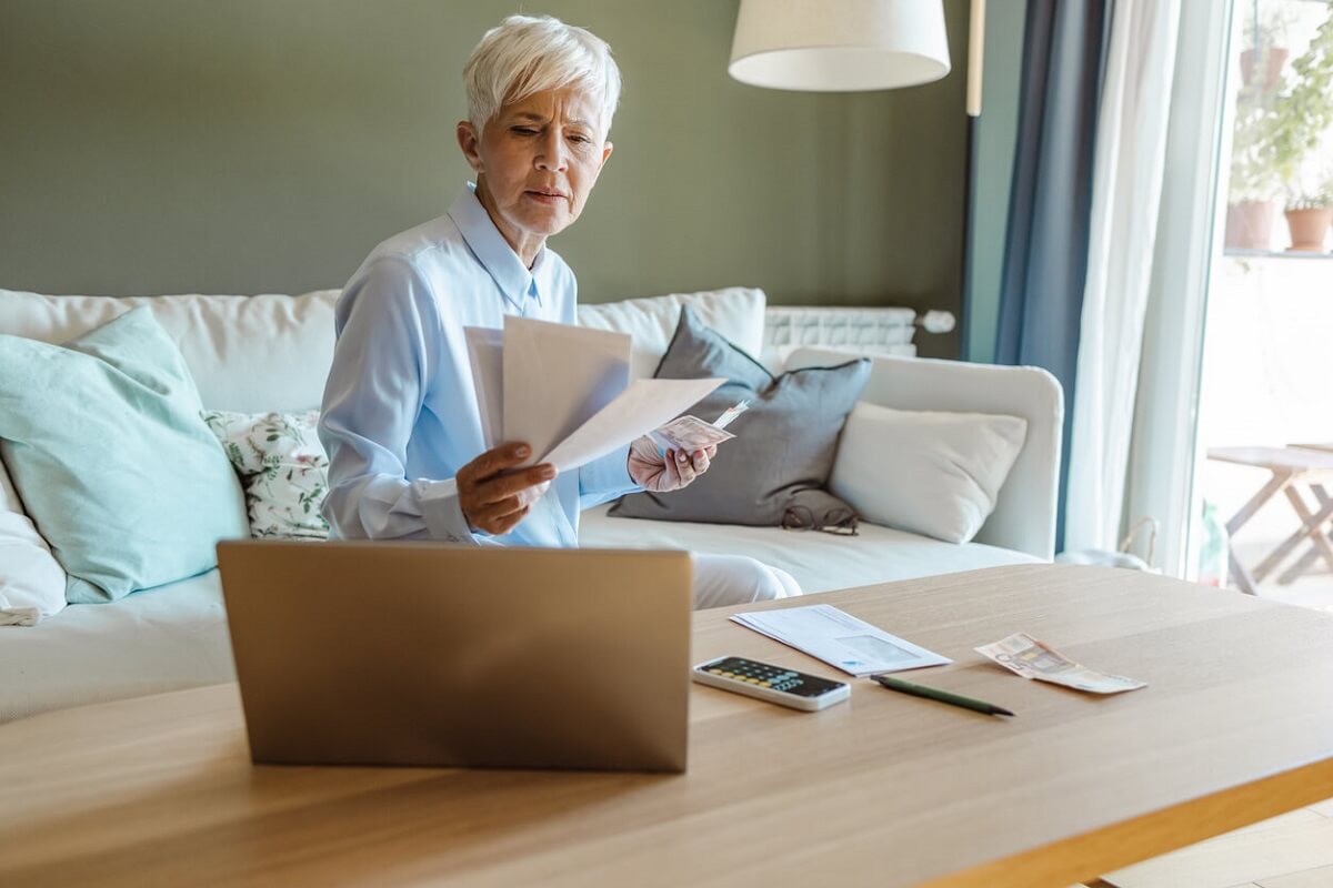 Une femme âgée en chemise bleue est assise dans le salon et examine des documents. 