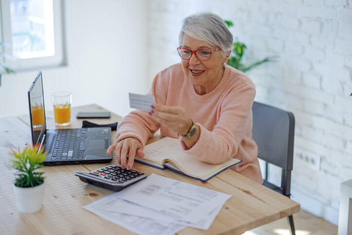 Femme âgée tenant une carte de crédit et utilisant un ordinateur portable tout en réglant des factures à la maison avec une carte de crédit.