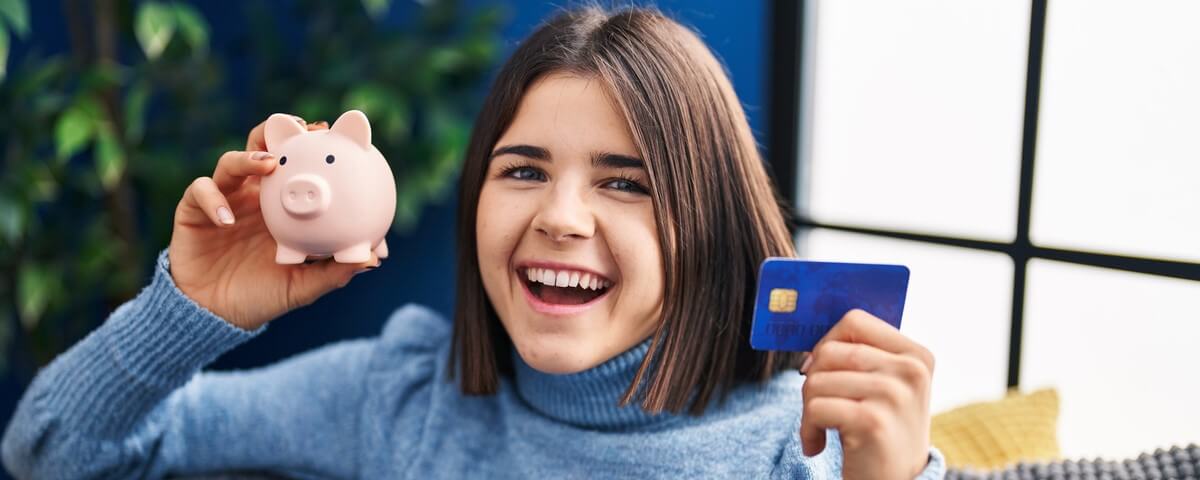 Jeune femme hispanique tenant une tirelire et une carte de crédit, souriant et riant à gorge déployée à cause d'une plaisanterie amusante et loufoque.