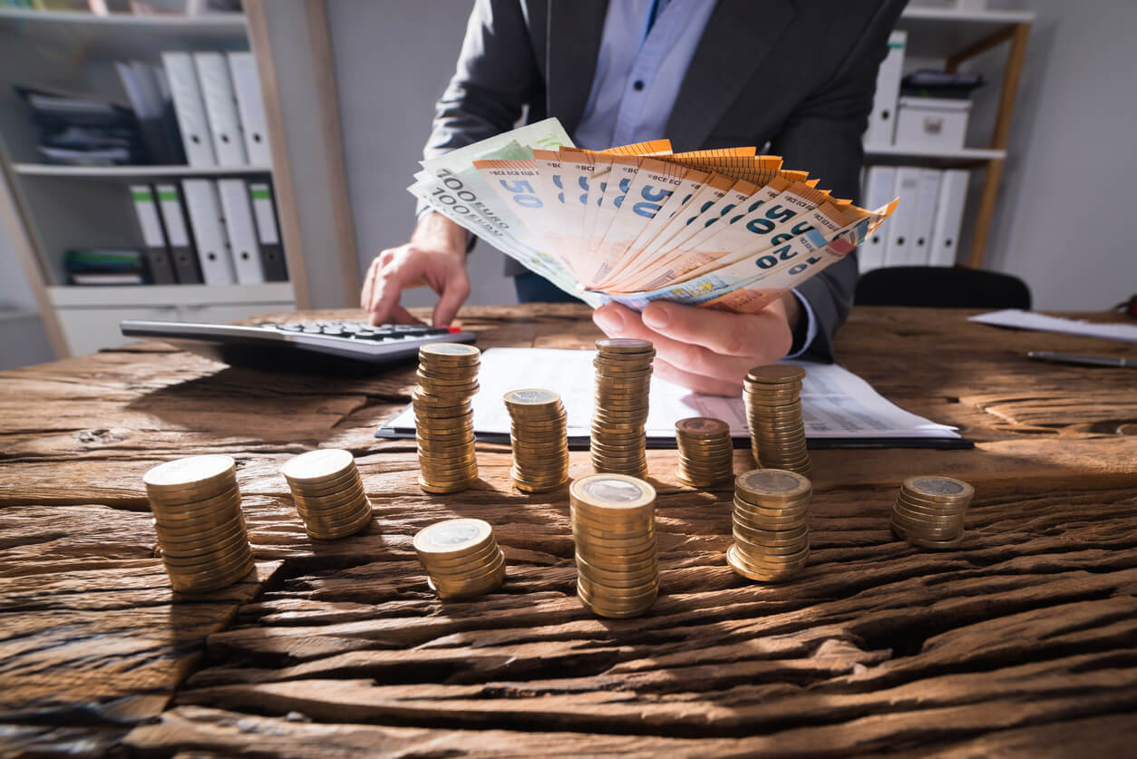 Homme d'affaires calculant des billets d'euros avec des pièces d'or empilées sur un bureau.