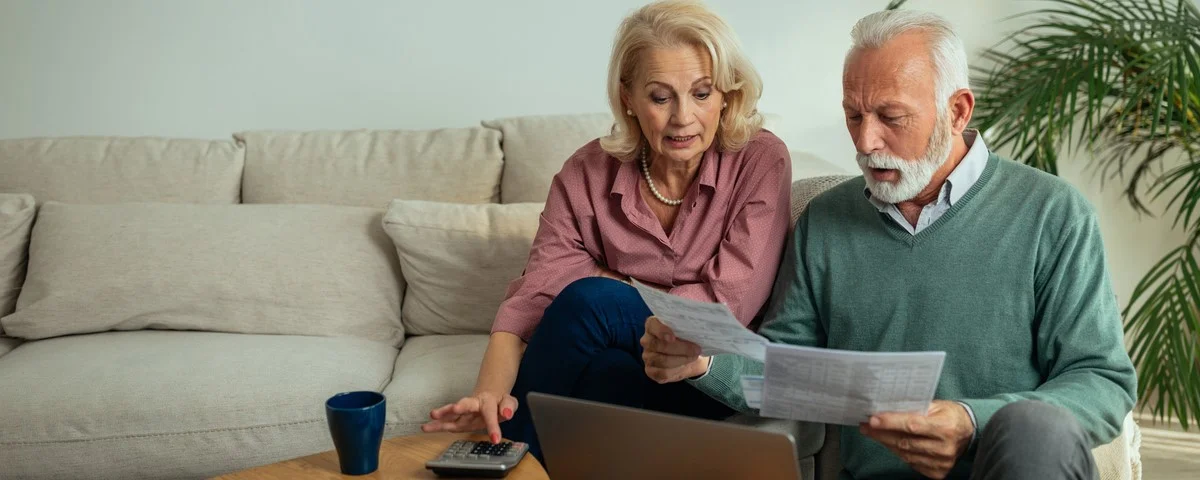 Couple de seniors utilisant un ordinateur portable, assis sur le canapé du salon, et examinant des documents à la maison.