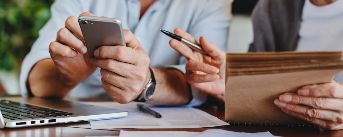 Fermez-vous vers le haut du couple âgé s’asseyant à la table dans le salon gèrent des factures de salaire de document de ménage utilisant le smartphone