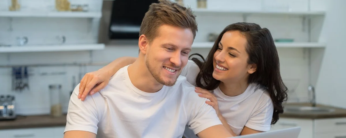 Taille haute d'un jeune couple souriant travaillant sur son ordinateur portable, assis dans la cuisine. Concept de relations.