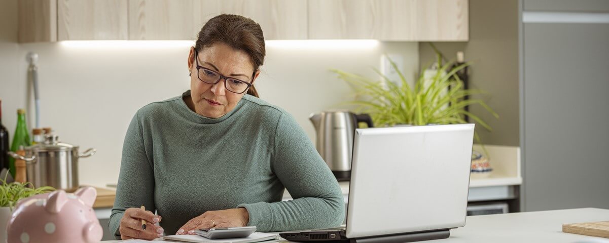 Portrait d'une femme hispanique d'âge mûr assise dans la cuisine en train d'analyser les finances de son foyer. Elle utilise un ordinateur portable et une calculatrice.