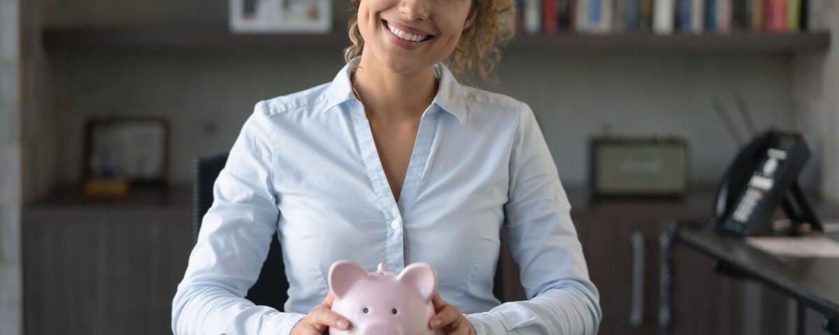Femme d'affaires heureuse au bureau économisant de l'argent dans une tirelire et regardant la caméra en souriant.