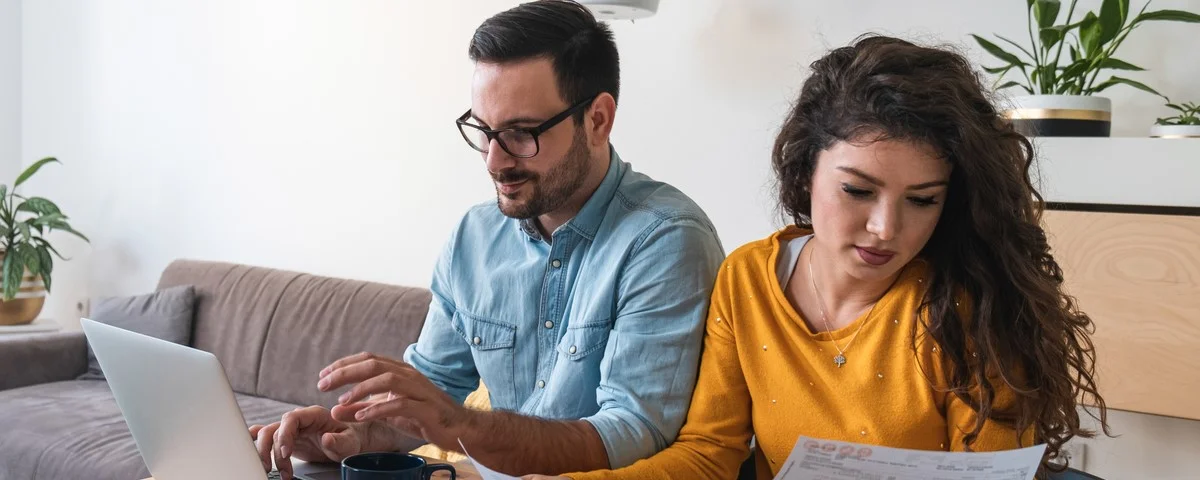 Mari et femme tenant des factures ou des impôts, calculant des dépenses ensemble, assis à un bureau et utilisant un ordinateur.