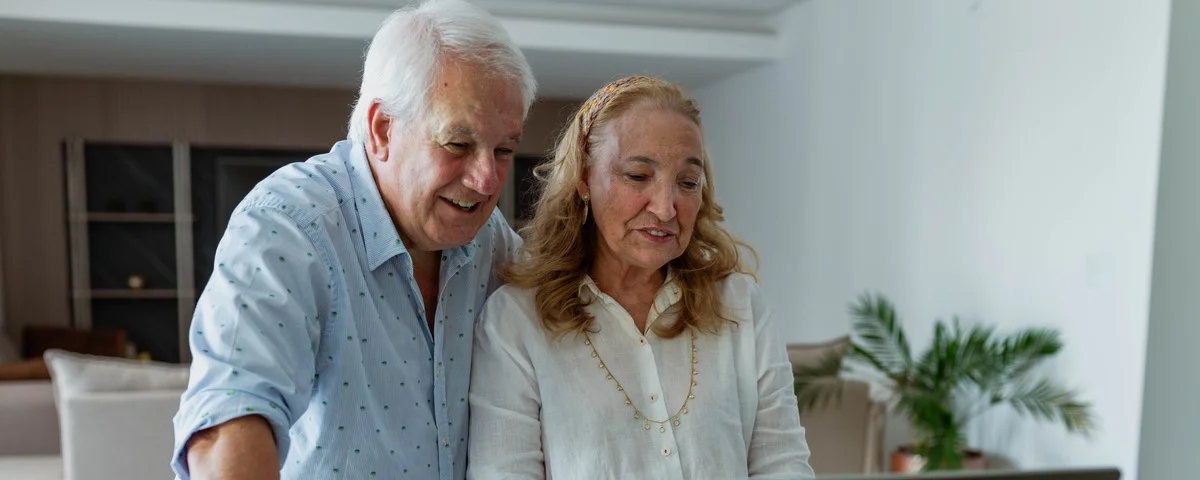 Couple de seniors souriants travaillant sur un ordinateur portable, debout à une table dans leur salon à la maison