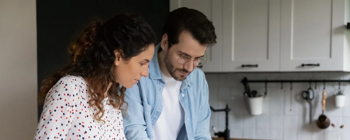 Homme portant des lunettes et femme vérifiant des documents financiers, planifiant un budget, debout à une table dans la cuisine, femme et mari calculant des factures.