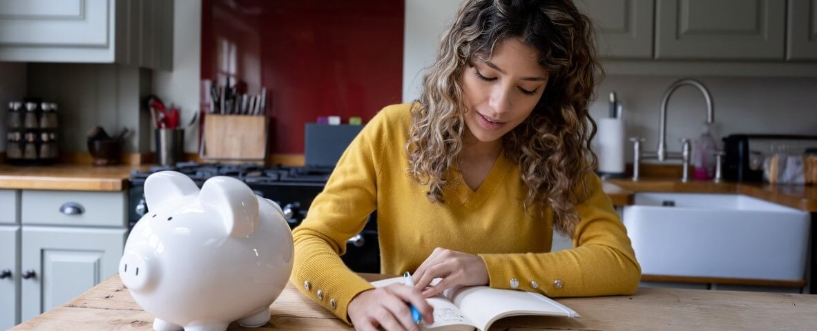 Jeune femme à la maison faisant son budget en écrivant sur un bloc-notes avec une tirelire à côté d'elle et des pièces de monnaie au premier plan.