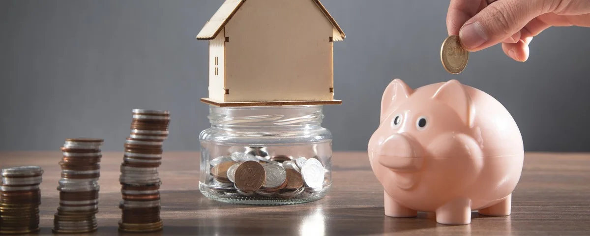 Main d'homme mettant une pièce dans une tirelire. Modèle de maison et pile de pièces sur la table