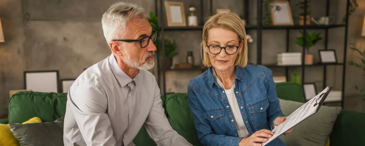 Un couple de seniors, mari et femme, travaillent ensemble depuis leur domicile. La femme tient un presse-papiers et discute de documents avec son mari.