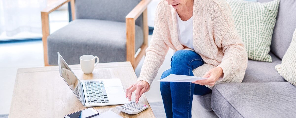 Prise de vue d'une femme âgée utilisant un ordinateur portable et une calculatrice tout en examinant des documents à la maison.
