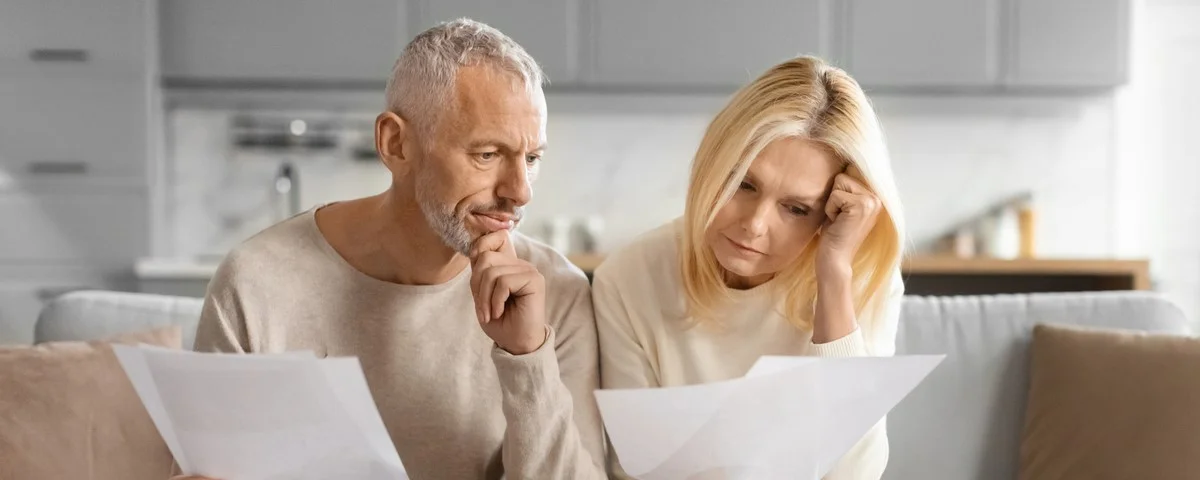 Couple mature regardant des documents avec inquiétude, assis sur un canapé avec une tablette et une tasse de café.