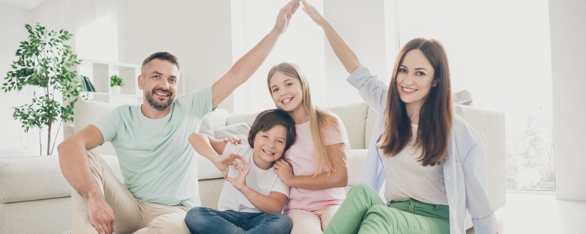Photo en grand format d'une famille joyeuse et chaleureuse — maman, papa et leurs deux enfants — en train de jouer, de dessiner un cœur sur le toit, assis par terre et portant des t-shirts chez eux
