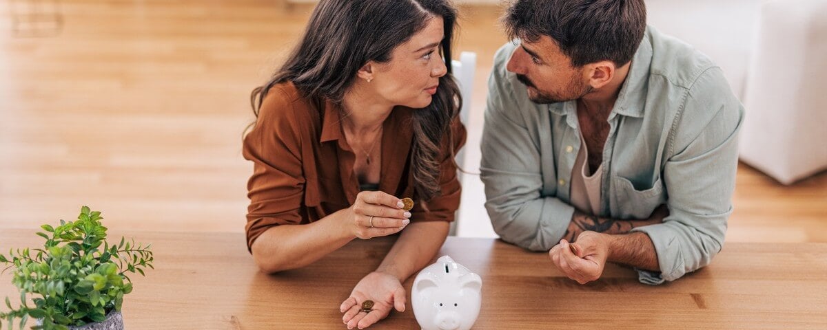 Un jeune couple assis à une table, inquiet de sa situation financière, comptant les dernières pièces et les derniers billets qui leur restent, avec une tirelire devant eux.