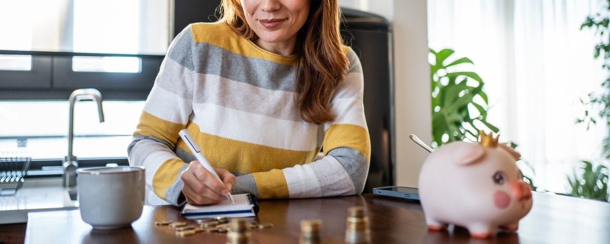 Une femme d'âge mûr assise à une table, prenant consciencieusement des notes sur les finances de son foyer tout en calculant ses économies et ses placements, entourée d'une tirelire, de pièces de monnaie et d'une tasse à café
