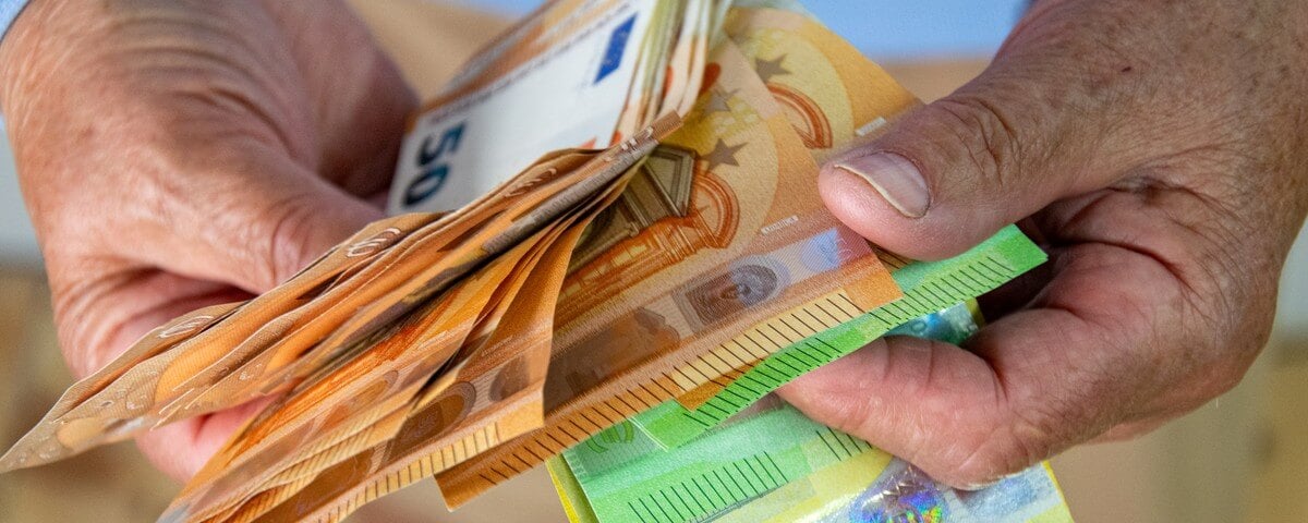Gros plan sur les mains d'une personne tenant une liasse de billets d'euro. Les billets sont disposés en éventail et la personne les tient entre le pouce et l'index.