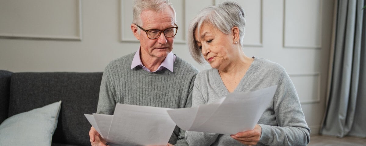 Un couple d’aînés d’âge mûr, tristes, fatigués et déçus, est assis devant des documents papier. Un homme et une femme d’âge mûr, l’air malheureux, lisent des factures, gèrent leurs finances bancaires, calculent leurs impôts, planifient leurs emprunts, leurs dettes et le versement de leur rente.