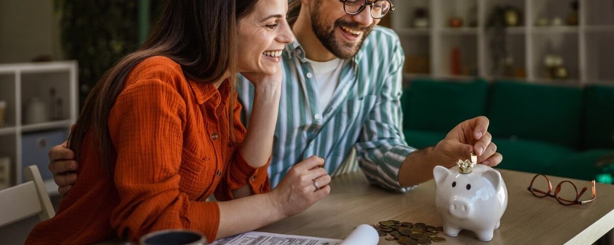 Un jeune couple heureux qui économise ensemble, en déposant des pièces dans une tirelire tout en discutant de leurs finances et en planifiant leurs futurs investissements, assis à une table à l'intérieur.