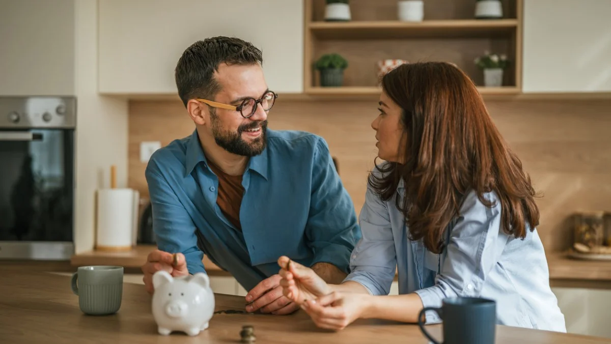 Un jeune couple déposant des pièces dans une tirelire posée sur le plan de travail de leur cuisine, souriant et discutant d'épargne à long terme, de gestion budgétaire et d'objectifs financiers dans le cadre chaleureux de leur foyer.