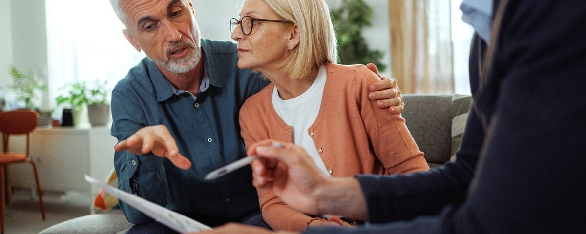 Un agent d'assurance présente des documents à un couple d'âge mûr chez eux, ils examinent les documents et discutent des différentes options de contrat dans une ambiance sereine et bienveillante.