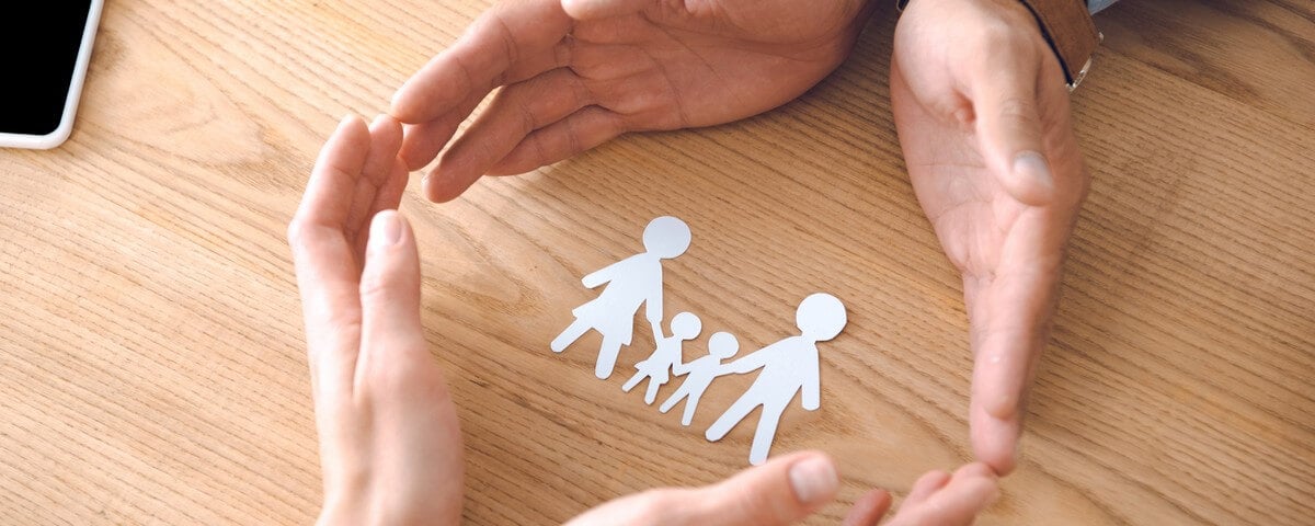 Vue partielle d'agents d'assurance et de mains féminines tenant une maquette en papier représentant une famille, posée sur un plateau de table en bois; concept d'assurance familiale